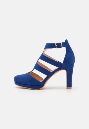 Chaussure à talon haut en daim bleu avec trois brides découpées et une boucle à la cheville sur fond blanc.