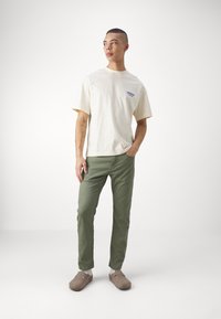 Cream short-sleeve t-shirt with blue logo, olive green slim-fit pants, and tan slip-on footwear. Smooth fabric and simple design.