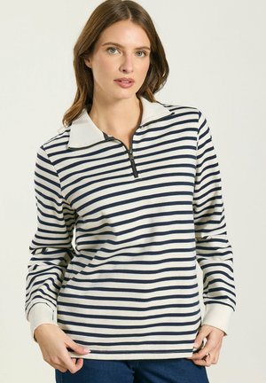 Femme portant un haut à manches longues à rayures blanches et marine avec un col zippé au quart, debout devant un fond clair uni.