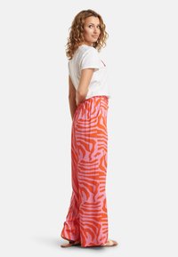 Lange, lichte broek in het roze met oranje zebra-stripes, voorzien van een elastische tailleband. Gecombineerd met een witte T-shirt met korte mouwen.