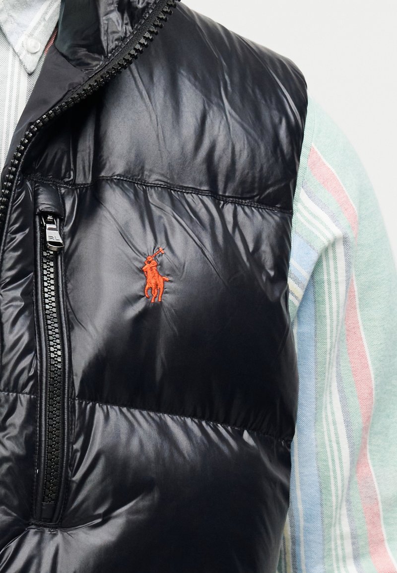 Veste sans manches matelassée noire avec une texture brillante et un col haut, dotée d'une poche zippée et d'un petit logo rouge brodé sur la poitrine.