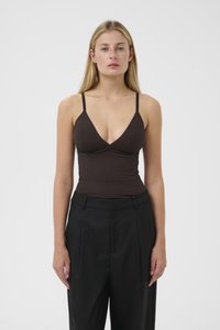 Donna con lunghi capelli biondi, vestita con un top senza maniche marrone scuro e pantaloni neri a vita alta, in piedi di fronte a uno sfondo bianco semplice.