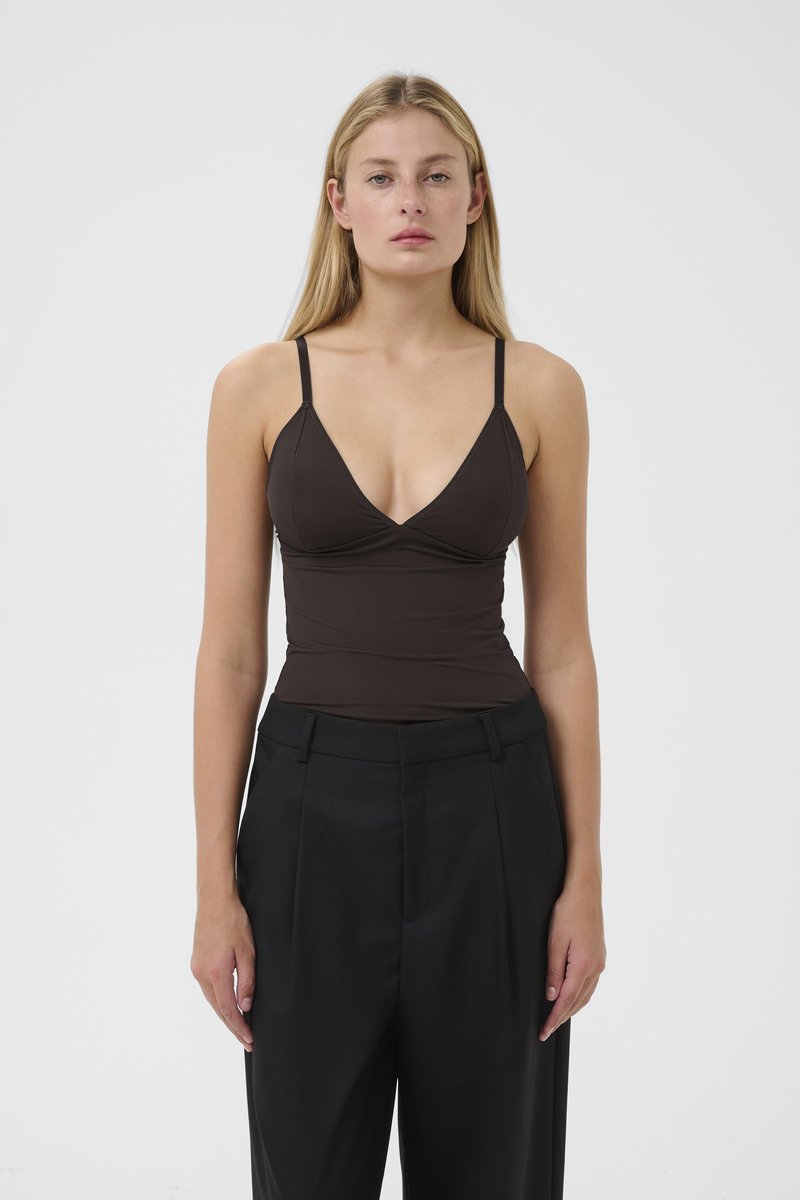Donna con lunghi capelli biondi, vestita con un top senza maniche marrone scuro e pantaloni neri a vita alta, in piedi di fronte a uno sfondo bianco semplice.