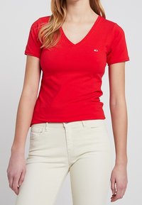 Rotes V-Ausschnitt-T-Shirt aus Baumwolle mit kurzen Ärmeln; verfügt über ein kleines Logo auf der Brust. Getragen mit hellen Jeans.