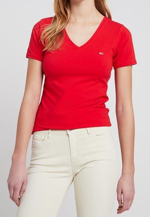 Maglietta rossa a V in cotone con maniche corte; presenta un piccolo logo sul petto. Indossata con jeans di colore chiaro.