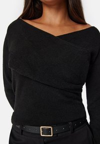 Zwarte gebreide trui met een asymmetrisch off-shoulder ontwerp, voorzien van een gevouwen paneel over de borst. Draagt met een zwarte riem en broek.