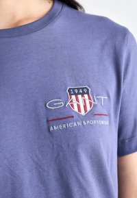 Violet medvilninė marškinėliai su siuvinėtu šarvuoti logotipu. Po logotipu balta spalva yra išsiuvinėti žodžiai „GANT“ ir „AMERICAN SPORTSWEAR“. 
