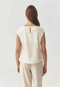 Blusa de color crema con mangas cortas, escote redondo y un detalle de abertura en la parte trasera. Tela suave con un corte holgado.