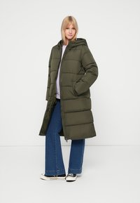 Hosszú, olívazöld steppelt kabát, kapucnival, elülső cipzárral, oldalsó zsebekkel és enyhén bővített fazonnal. Farmernadrág és sportcipő fölött viselve.