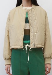 Giacca bomber trapuntata beige con chiusura a zip, polsini a costine e design cropped, abbinata a un maglione a righe in maglia e pantaloni verdi.