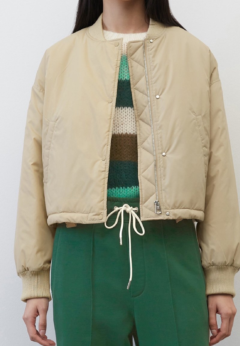 Giacca bomber trapuntata beige con chiusura a zip, polsini a costine e design cropped, abbinata a un maglione a righe in maglia e pantaloni verdi.