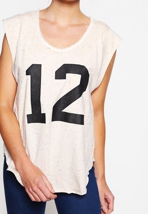White sleeveless top with black number 12, worn by person with one hand holding the hem, paired with dark blue pants.