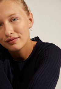 Pull en maille côtelée bleu marine avec un col à volants, présentant une texture lisse. Le mannequin porte une petite boucle d'oreille en or et a un maquillage naturel.