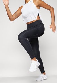 Leggings noirs avec une taille haute, présentant des coutures contrastantes, associés à un haut blanc court sans manches. Chaussures de sport blanches.
