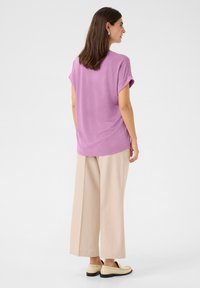 Violeta īsajā piedurknē augšdaļa ar sarullētām piedurknēm, apvienota ar bēšiem platām biksēm un gaišiem loafers, skatīta no aizmugures.