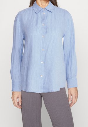 Frau trägt ein hellblaues, langärmliges Hemd mit dezenten vertikalen Streifen und graue gerippte Hosen.