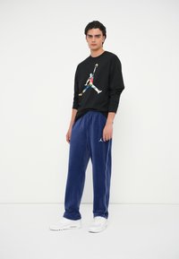 Μαύρη μπλούζα με μακριά μανίκια και πολύχρωμο γραφικό σχέδιο στο μπροστά μέρος, σε συνδυασμό με μπλε βελούδινους παντελόνες που έχουν λευκή πλευρική ρίγα και λογότυπο.