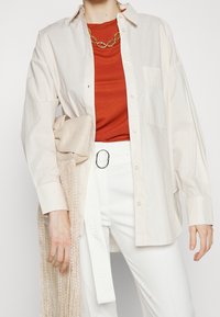 Lichtbeige overhemd met lange mouwen en een knoopsluiting, gecombineerd met een oranje top en witte broeken. Accessoires zijn een kettinghalsketting en een geweven tas.