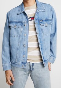 Uomo che indossa una giacca di jeans azzurro chiaro sopra una camicia a righe beige e bianca della Tommy Jeans e jeans azzurri, in piedi con una mano in tasca.
