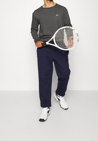 Long-sleeve gray shirt with a small green logo, paired with navy sweatpants. The model holds a white tennis racket and wears white sneakers.