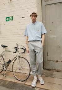 Camisa azul clara de mangas curtas com textura, calças de moletão cinzas, ténis brancos, em pé ao lado de uma bicicleta, contra uma parede de tijolos e uma porta.