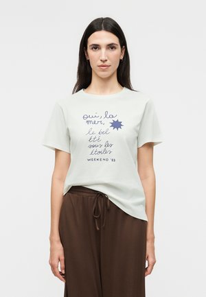 Donna con capelli lunghi e scuri che indossa una maglietta bianca a maniche corte con scritta in francese e pantaloni marroni su uno sfondo semplice.