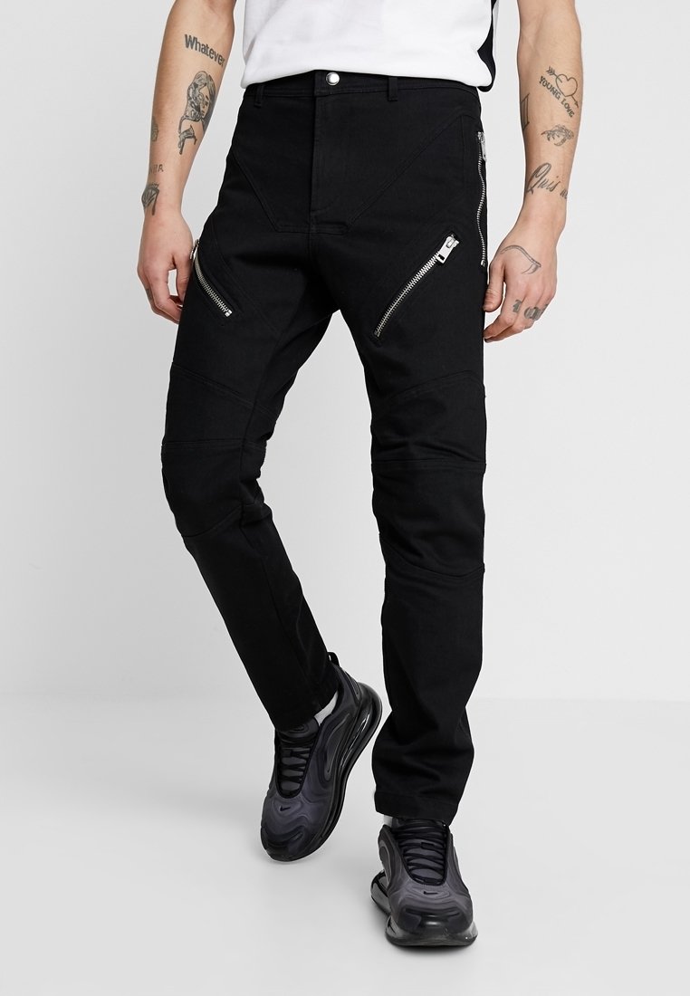 Homme portant un pantalon noir slim avec des poches zippées diagonales argentées et des baskets noires, debout jambes croisées.