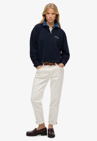 Pull marin avec un col bleu, logo brodé, associé à un pantalon blanc et des mocassins foncés, avec des manches retroussées et une ceinture tressée.