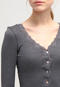 Grijze geribbelde longsleeve top met een kantafwerking bij de V-hals en kleine knopen aan de voorkant. Sluit nauw aan met een gladde textuur.