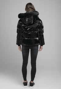 Czarna kurtka puchowa z kapturem z sztucznego futra, błyszcząca tekstura, długie rękawy i poziome pikowane sekcje, w połączeniu z czarnymi obcisłymi dżinsami i trampkami.