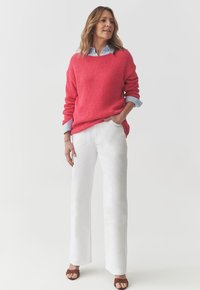 Maglione lavorato a maglia corallo con texture a coste, indossato sopra una camicia azzurro chiaro con colletto, abbinato a pantaloni bianchi a gamba larga e sandali marroni.