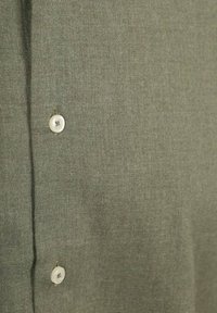 Olijfgroene stof met een textuurafwerking, voorzien van een rij witte knopen. De stof lijkt zacht, met een subtiel weefpatroon.