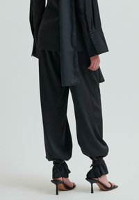 Czarny satynowy zestaw składający się z bluzy z długim rękawem i luźnych spodni z marszczonymi kostkami oraz spiczastych sandałów na wysokim obcasie.