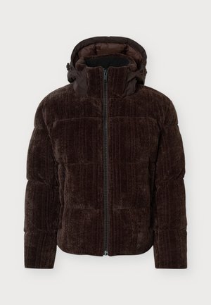 Un homme porte une doudoune à capuche marron foncé, un jean gris et des bottes noires sur un fond clair uni.