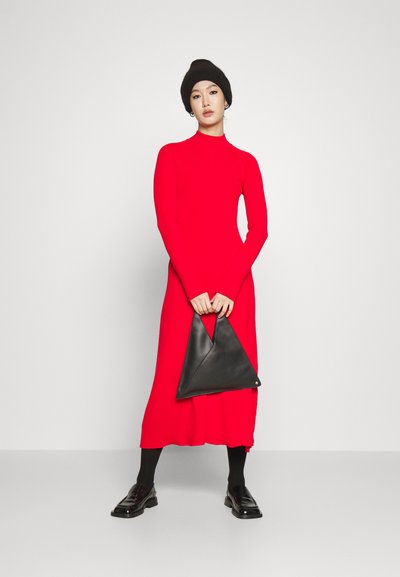 Robe bodycon rouge à manches longues avec un col montant ; associée à un sac à main en cuir noir en forme de triangle et des chaussures noires, portée avec des collants noirs.