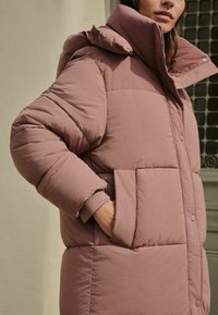 Palton captusit în nuanță moale de mauve, cu guler înalt, nasturi cu sistem de prindere și două buzunare frontale, având o textură netedă.