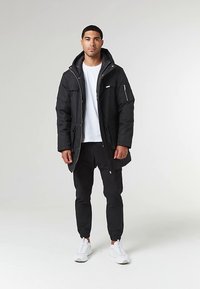 Jachetă neagră lungă, de tip puffer, cu glugă, închidere cu fermoar și buzunare laterale, asortată cu un tricou alb și pantaloni negri conici. Adidași albi.