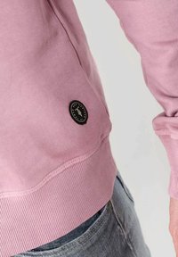 Felpa rosa con orlo e polsini a coste, caratterizzata da un patch circolare nero sul lato inferiore. Tessuto morbido con una texture liscia.