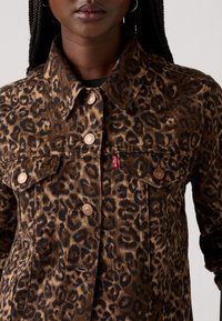 Veste en denim à motif léopard avec quincaillerie en bronze, fermeture à boutons sur le devant, col pointu et poches poitrine doubles. Textures marron clair et noir.