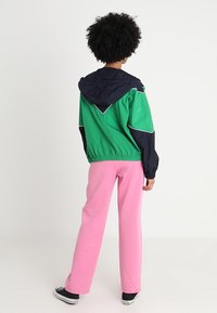 Person med krøllet hår iført en grøn og marineblå hættetrøje, lyserøde bukser og sorte sneakers, stående med ryggen til på en hvid baggrund.