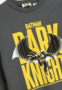 Sudadera gris con un gran gráfico amarillo que dice "BATMAN DARK KNIGHT" con una silueta de murciélago negra y una figura detallada de Batman en vuelo.