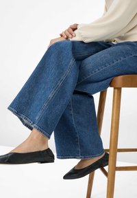 Jeans di denim blu a gamba larga con orli sfilacciati, appoggiati su uno sgabello di legno, abbinati a scarpe flat nere. Il tessuto ha una classica texture denim.