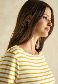 Gelb-weiß gestreifte Boot-Cut-Bluse mit strukturiertem Strick. Das Modell mit langen Haaren zeigt sich von links vor einem neutralen Hintergrund.