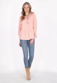 Pænt farvet, tekstureret bluse i fersken med en samlet peplum talje og bindebånd ved halsen, parret med blå jeans og brune cowboy støvler.