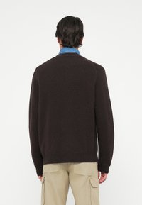 Brun stickad tröja med rund hals och långa ärmar, med en ribbad nederkant och manschetter, bärs över en jeansskjorta.