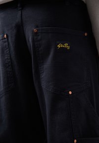 Ciemnogranatowe spodnie cargo z dużymi kieszeniami i miedzianymi elementami ozdobnymi. Na tylnej kieszeni znajduje się żółta naszywka z logo. Wykonane z wytrzymałej tkaniny.