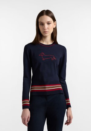 Donna che indossa un maglione blu navy con strisce rosse e beige sui polsini e sull'orlo, con il profilo di un bassotto rosso sul petto, in piedi.