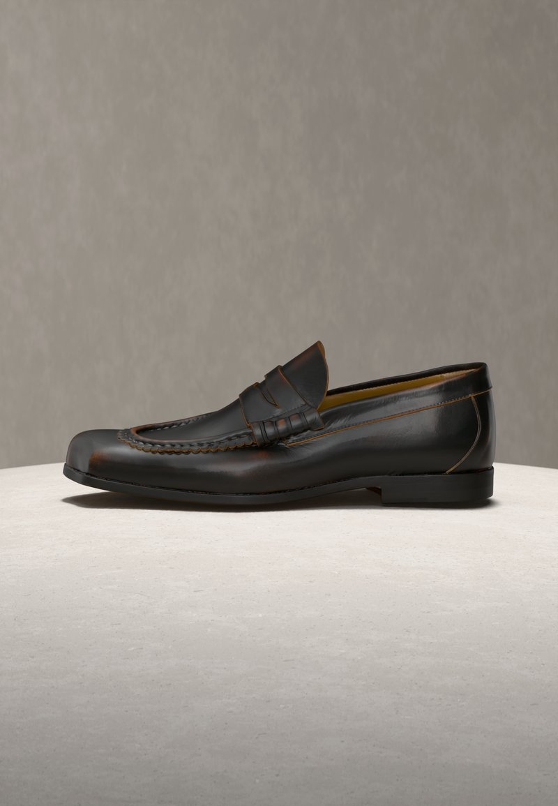 Black leather penny loafer with stitched detailing on a light surface and plain beige background.