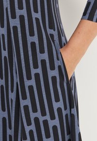 Sininen mekko, jossa on geometrinen musta kuvio, lyhyet hihat ja sivutaskut. Valmistettu pehmeästä, kevyestä kankaasta, jossa on rento istuvuus.