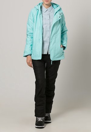 Světlomodrá nepromokavá bunda s kapucí, předními kapsami a zapínáním na zip, ve spojení s černými kalhotami a černými teniskami s bílými tkaničkami.